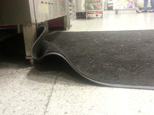 mat that is loose and not secured to the ground. It is raised off the ground and forms a lip. Next to a soda machine 