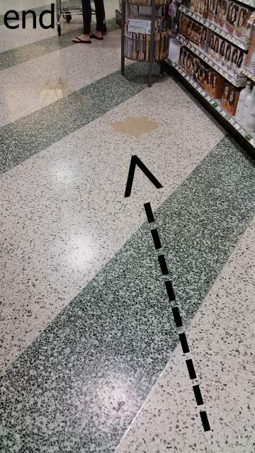 Arrow pointing to beige puddle of liquid on the floor of a supermarket. 