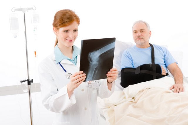 treating doctor at hospital reviewing x-ray. Man in bed with sling on his arm.