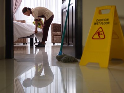 Lady making bed at hotel. Mop on the floor and yellow caution wet floor sign.